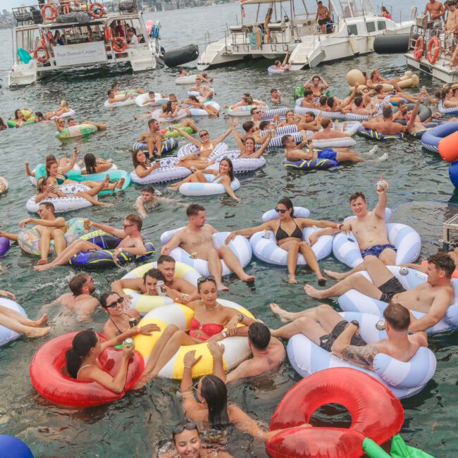 A large group of people relax on colorful inflatable tubes and rafts in the water near boats, enjoying a lively social gathering under a cloudy sky.