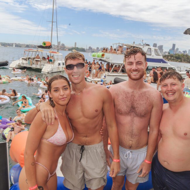 Four young adults in swimwear stand together smiling at a lively boat party, with boats, people swimming on inflatables, and a city skyline visible in the background under a partly cloudy sky.