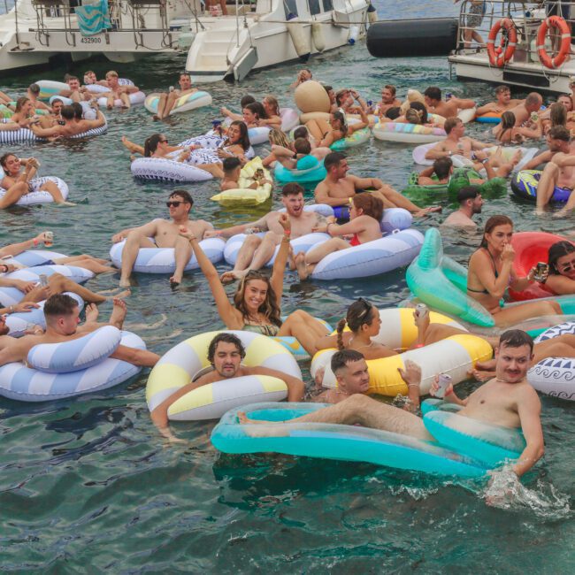 A large group of people relax and socialize on inflatable pool floats in the water near anchored boats, enjoying a lively gathering on a sunny day.