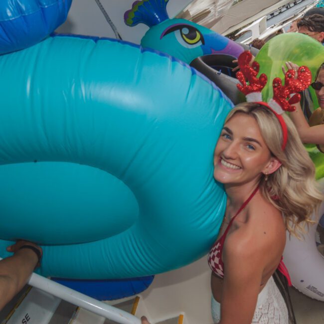 A smiling woman in a red swimsuit and reindeer antlers carries a large blue inflatable ring on a boat. Other people in swimsuits and inflatables are nearby, with water and boats visible in the background.