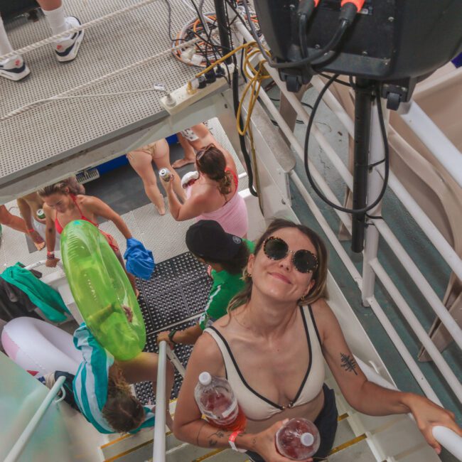 A woman in sunglasses and a bikini holds drinks while standing on stairs at a pool party. Below her, people in swimsuits and beachwear socialize and carry floaties. The scene is energetic and summery.