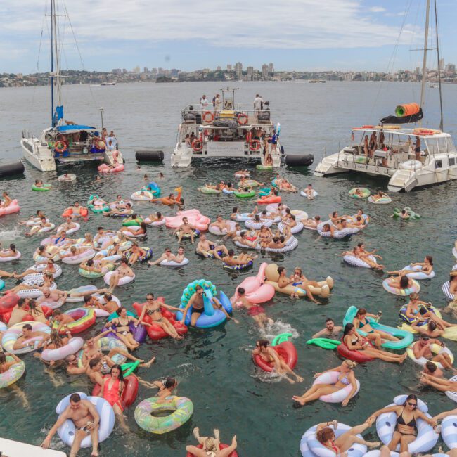 A large group of people relax and socialize on colorful inflatable pool floats in the water near several anchored boats on a sunny day. The scene is festive and crowded, with many swimmers enjoying the event.