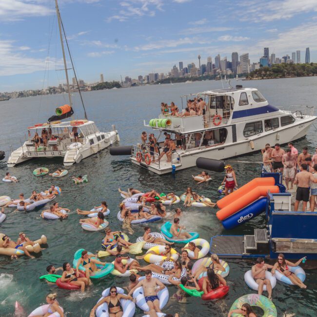 A large group of people party on boats and colorful inflatable floats in the water. The Sydney skyline and Harbour Bridge are visible in the background on a sunny day. Signs and decorations give a festive atmosphere.