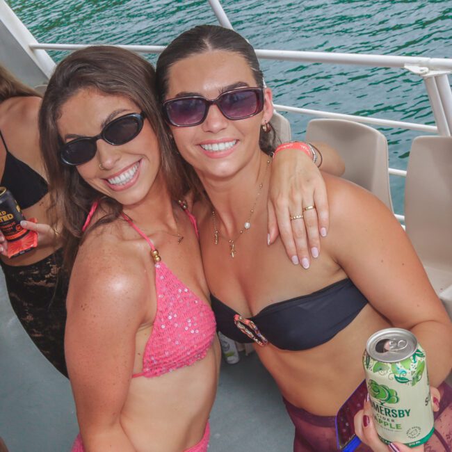 Two women in swimsuits, wearing sunglasses and smiling, stand closely together on a boat, each holding a canned drink. The background shows blue-green water and trees on a sunny day.