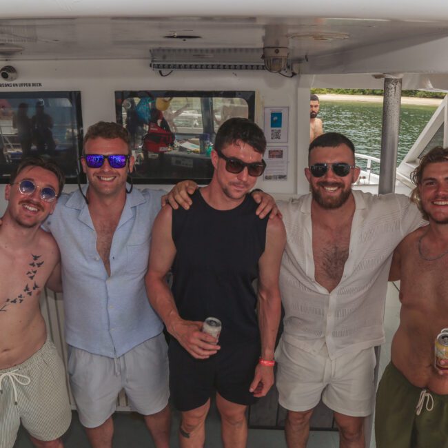 Five men in casual summer clothes stand together on a boat, smiling and holding drinks. The water and deck are visible in the background, and a “Yacht Social” logo is in the bottom right corner.
