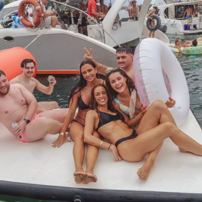 A group of young adults in swimsuits smile and pose together on a floating mat in the water near boats, surrounded by other people enjoying a lively pool party.