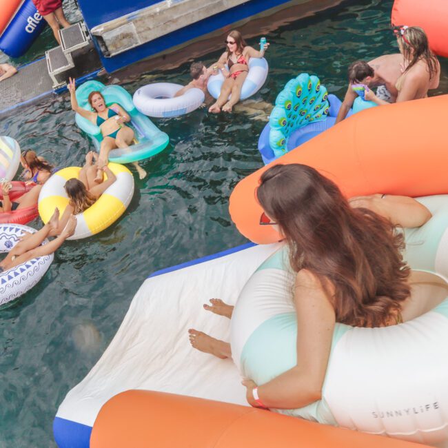 A group of people relax on colorful inflatable floats in a lake, chatting and enjoying the water. One woman slides down an inflatable slide toward the group. The scene is lively and summery.
