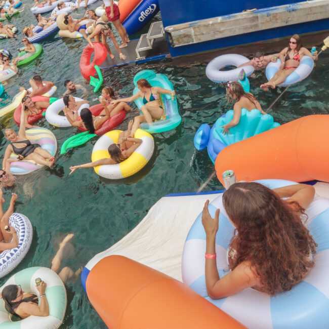 A large group of people on colorful inflatable floats relax and socialize in the water near boats on a sunny day. Some hold drinks, while others lounge, enjoying a lively, festive atmosphere.