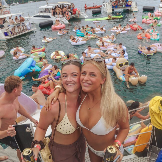 Two women in swimsuits smile and pose on a boat with drinks in hand, surrounded by people on pool floats and nearby yachts at a lively outdoor water party. The atmosphere is festive and summery.