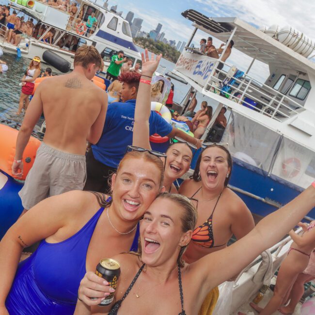 A group of young people in swimsuits smile and pose with drinks on a crowded boat, surrounded by others on inflatables and nearby boats on a sunny day, with a city skyline in the background.