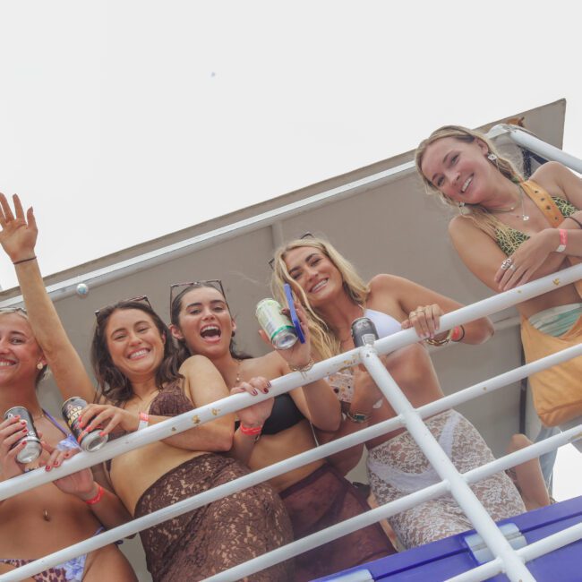 Five young women in swimsuits stand on a boat deck, smiling and holding drinks, enjoying a sunny day. Some are waving or leaning on the railing. The sky is bright and overexposed in the background.