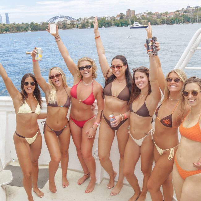 Seven women in bikinis stand on a boat, smiling and raising drinks in celebration. A city skyline and bridge are visible in the background across the water on a sunny day.