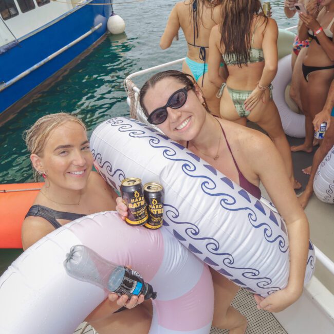 Two smiling women in swimsuits holding inflatable pool rings and drinks pose on a boat. Other people in swimwear stand nearby, and a blue boat is anchored in the background on clear water.