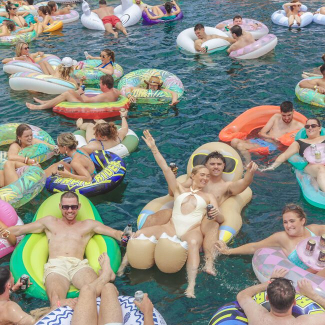 A large group of people relax on colorful inflatable floats, chatting and holding drinks, in a crowded, lively pool under bright sunlight. Many are laughing, enjoying summer attire, and the scene feels festive and joyful.