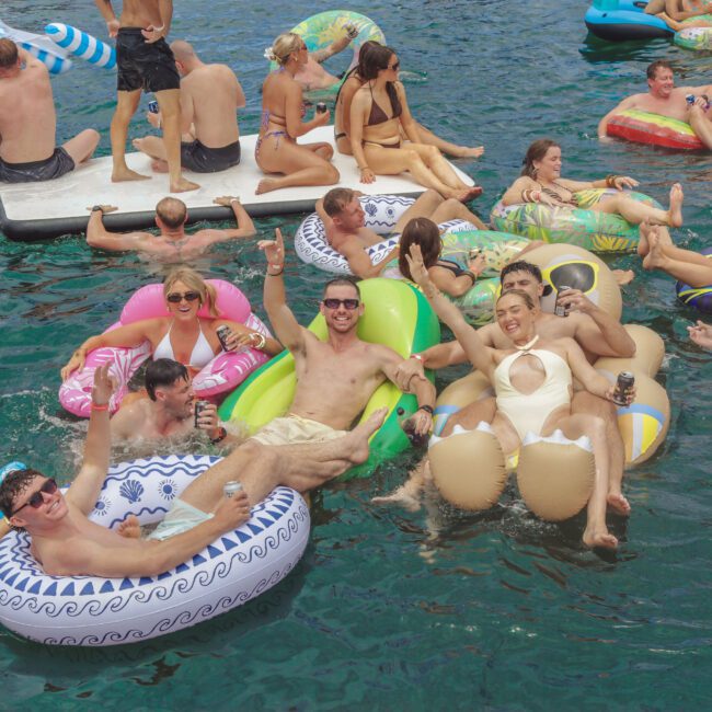 A group of people relax and enjoy drinks on colorful inflatable pool floats in a lake, surrounded by others on floats and a floating platform, all under bright daylight. The atmosphere is lively and festive.