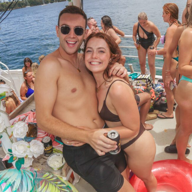 A smiling man and woman in swimsuits pose together on a crowded boat party with people in swimwear, drinks in hand, and water visible in the background.