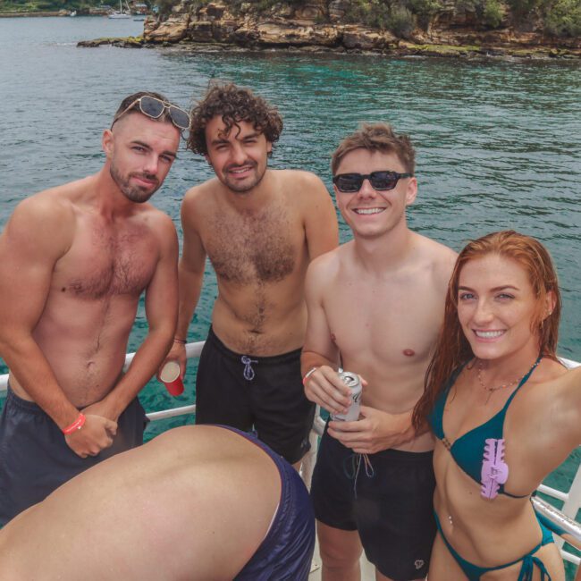 Four young adults in swimwear, three men and one woman, smile and pose on a boat with water and rocky green cliffs in the background. The woman raises her arm, and some people are partially visible around them.
