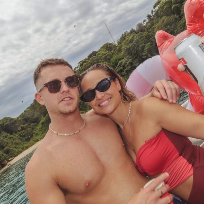 A man and woman in swimsuits and sunglasses relax on an inflatable float on the water, smiling and holding drinks with a green, tree-lined shoreline in the background.