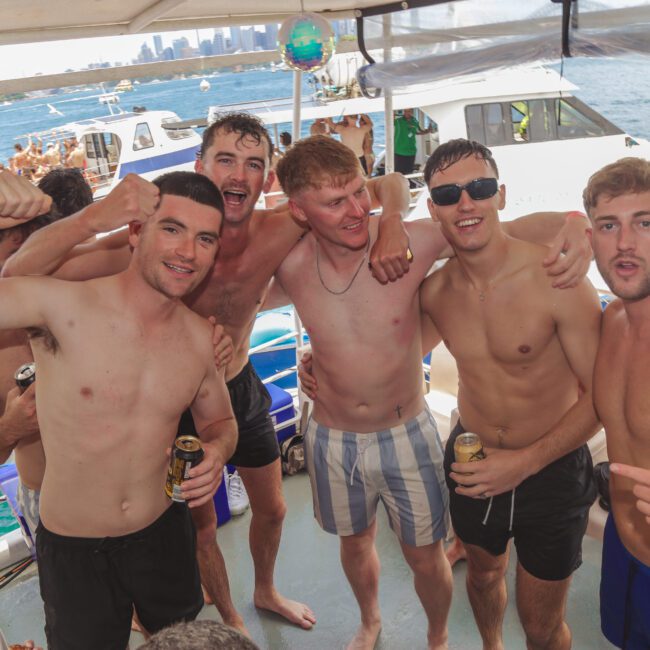 A group of five young men, shirtless and smiling, pose together on a boat with drinks in hand. Other people, boats, and water floaties are visible in the background under a sunny sky.