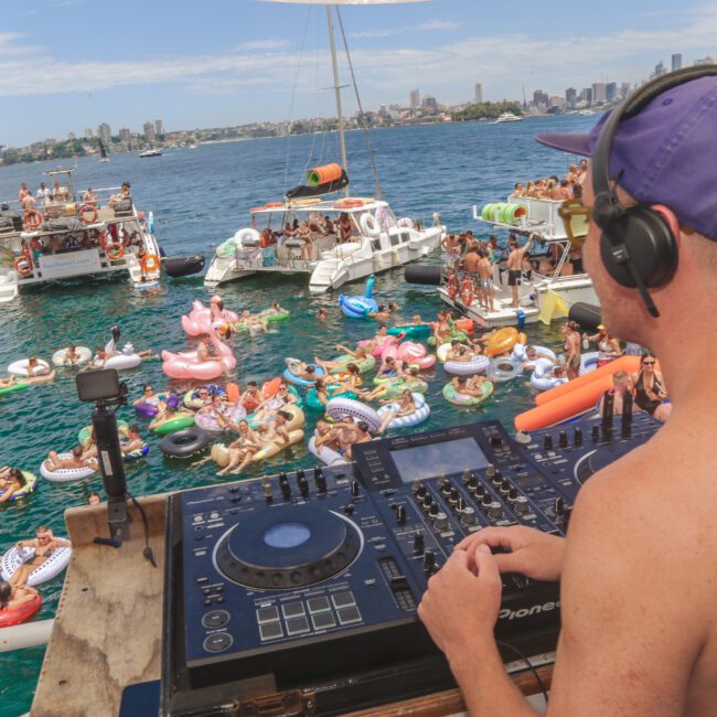 A DJ wearing headphones and a purple cap plays music on a boat, overlooking a lively crowd on inflatable pool floats and boats in the water, with a city skyline in the background.