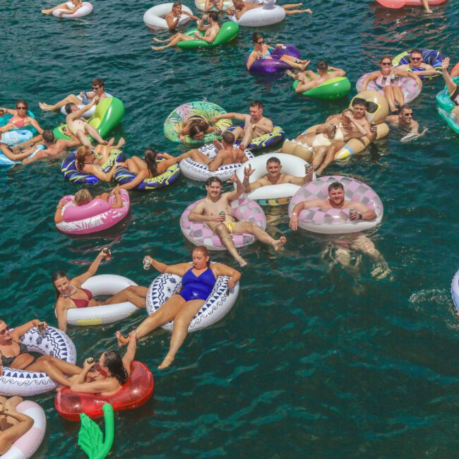 A large group of people relax on colorful inflatable pool floats in blue-green water, smiling and enjoying a sunny day. Some are lounging in groups, while others float alone. The Yacht Social Club logo appears in the bottom right corner.