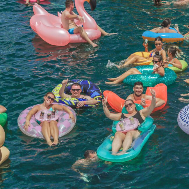 A group of people relax on colorful inflatable pool floats in blue water, smiling, holding drinks, and enjoying a sunny day. Some floaties are shaped like animals or pool rings. The scene is lively and festive.