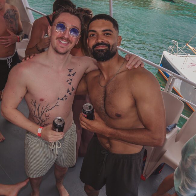 Two smiling men in swimwear stand on a boat deck with drinks in hand, surrounded by people in swimsuits. Blue water and another boat are visible in the background. The atmosphere is lively and relaxed.