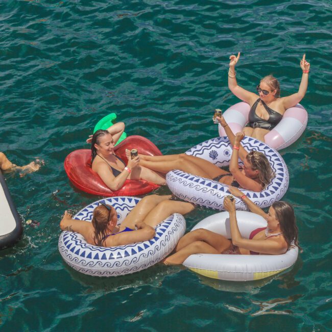 A group of women in swimsuits relax on inflatable floats and a dock, smiling and raising drinks, enjoying the sun on clear blue water. The scene looks festive and joyful.