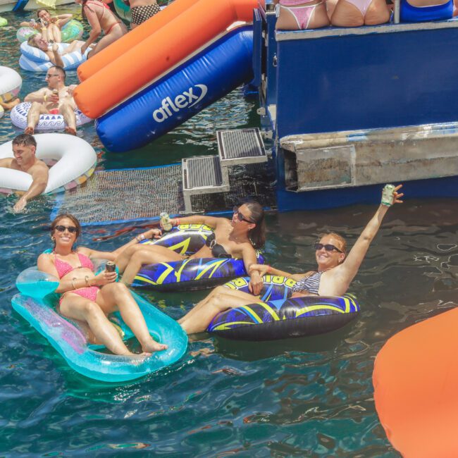 People relaxing on colorful inflatable floaties in the water near a docked boat. Three women in sunglasses and swimsuits smile and wave at the camera. Other people are floating and socializing nearby.