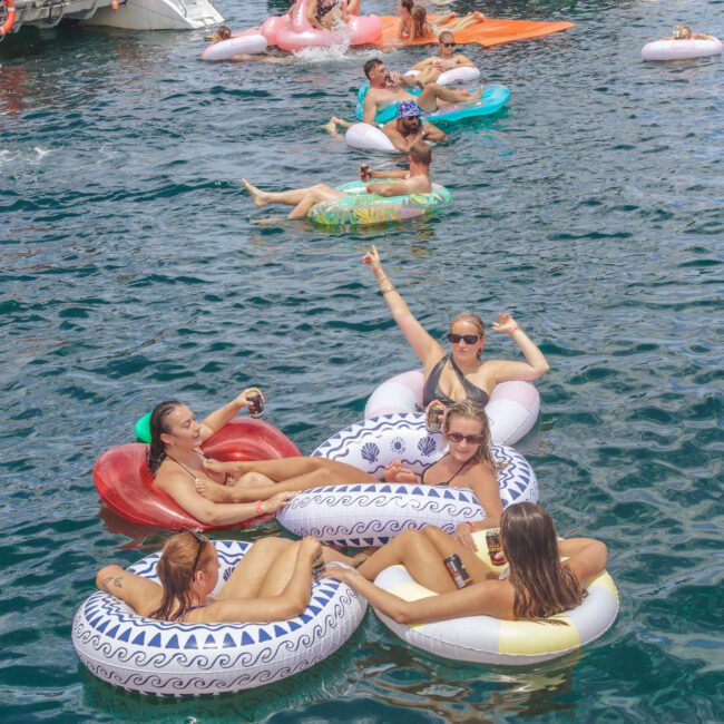 Groups of people relaxing on colorful inflatable rings in deep blue water, enjoying a sunny day. Some are smiling and posing for photos, while others float nearby. A small logo reads "Yacht Social Club" in the corner.