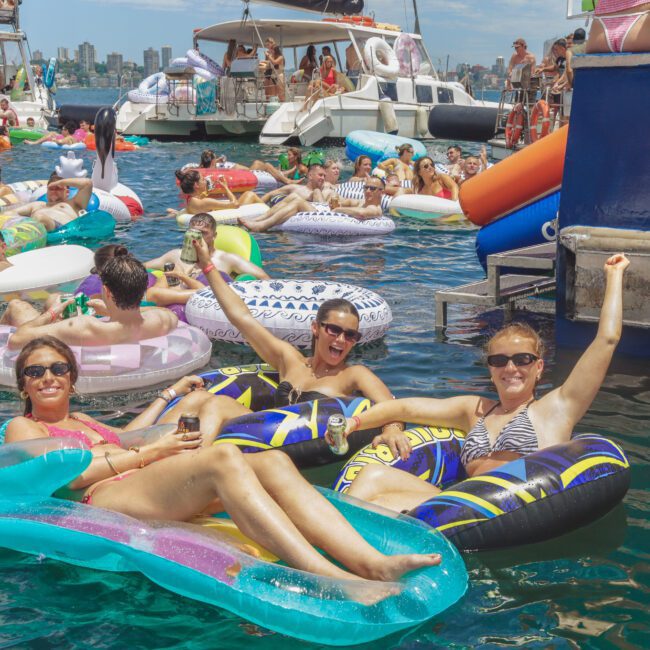 A lively crowd floats on colorful inflatable tubes in a marina, smiling and celebrating under sunny skies, with yachts and city buildings in the background. Three women in sunglasses cheer and relax in the foreground.
