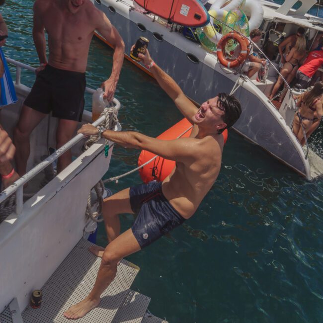 A man in swim trunks, laughing and holding a drink, leans back dramatically on boat steps while another man steadies him. Several people relax on a nearby catamaran in the water, with inflatable pool toys visible.