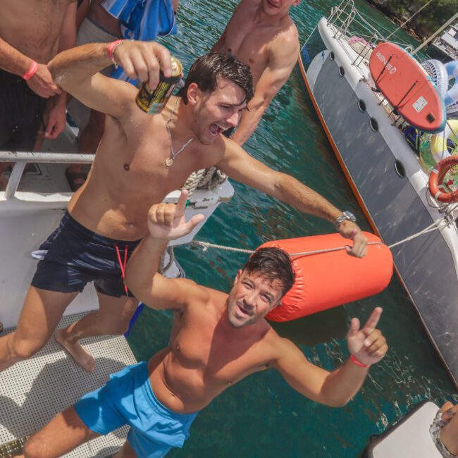 A group of smiling, shirtless men on a boat enjoy a sunny day, with one man holding a drink and another giving a thumbs up. A yacht and lush green trees are visible in the background over turquoise water.