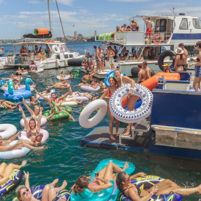 A lively scene of people in swimsuits relaxing on colorful inflatable tubes and floaties in the water near docked boats during a sunny day, with a city skyline visible in the background.