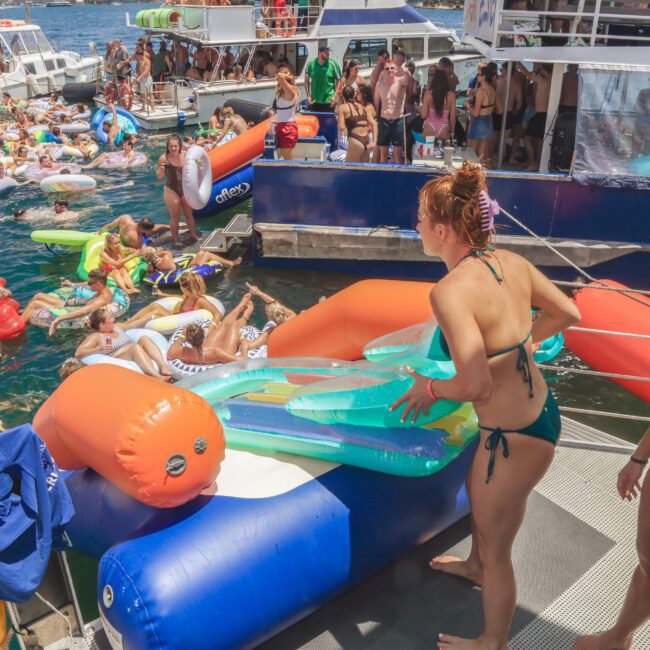 People relaxing on colorful inflatables in the water near boats, with others standing on a dock. Two women in swimsuits are in the foreground, one holding a float, and it is a sunny day.