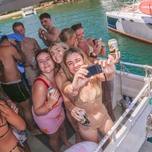 A group of young adults in swimsuits smile and pose for a selfie on a crowded boat, holding drinks, with water and another boat visible in the background on a sunny day.