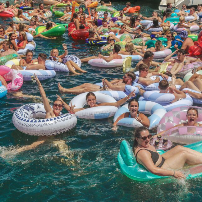 A large group of people relax on colorful inflatable floats in the water, enjoying a sunny day. Many are smiling, waving, or holding drinks, creating a lively, festive atmosphere.