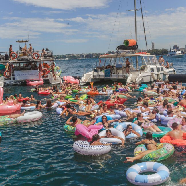 A large group of people relax on colorful inflatable floats in the water near boats, enjoying a sunny day with city buildings visible in the background.