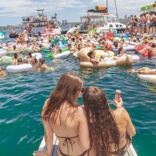 A large group of people enjoying a lively party on floaties and boats in the water under a sunny sky; two women in swimsuits sit at the edge with drinks, looking toward the crowd.
