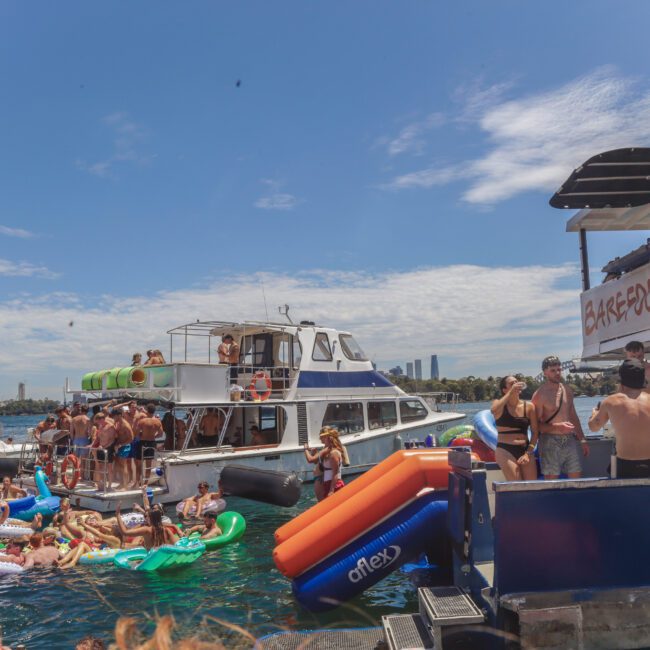 A lively boat party on the water with many people on yachts and inflatables, swimming, and socializing under a sunny sky. "BAREFOOT BLUE" is written on one boat. City buildings and blue sky are visible in the background.