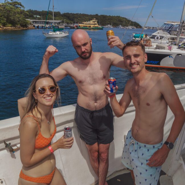 Three people in swimwear pose and smile on a yacht, holding drinks. The man in the center flexes his arms. The ocean and other boats are visible in the background under a clear sky.