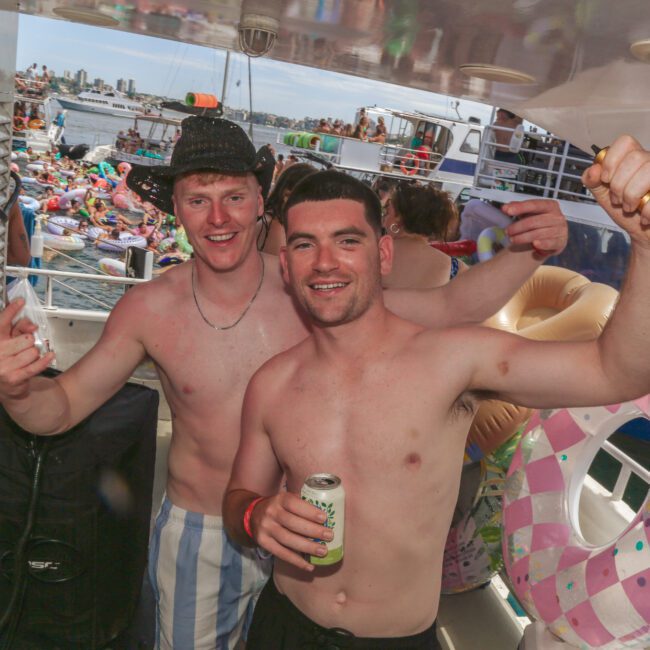 Two smiling shirtless men pose on a boat, one holding a drink. Colorful pool floats and other boats with crowds of people are visible in the background on the water.