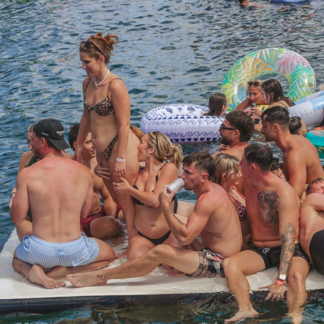 A group of young adults in swimwear gather closely on a floating mattress in a lake, surrounded by inflatable pool floats and others enjoying the water on a sunny day.