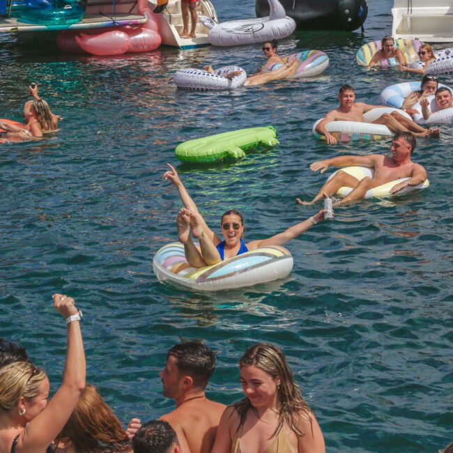 A group of people enjoy a pool party, floating on colorful inflatable rafts and swimming in clear blue water near boats. Most are wearing swimsuits, smiling, and having fun in the sun.