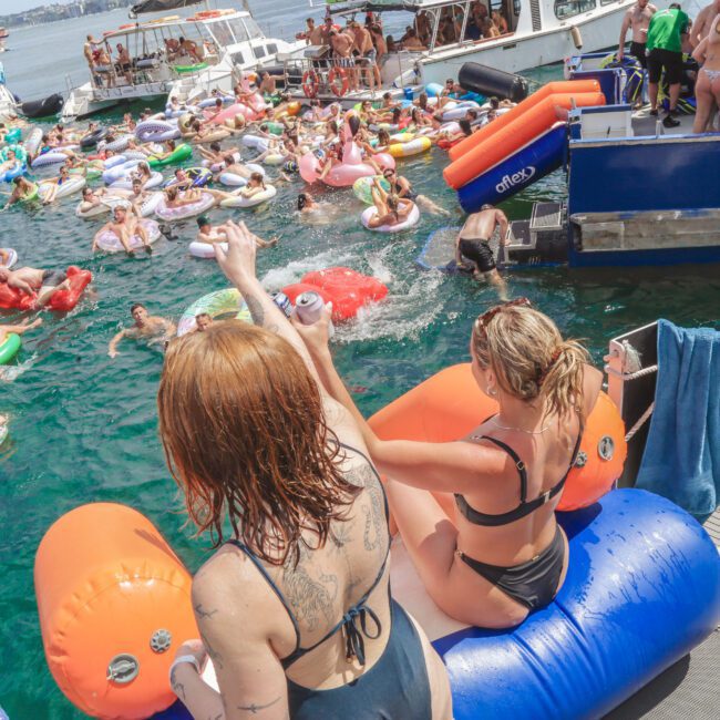 Two women in swimsuits sit on inflatable loungers at the edge of a dock, waving to a large crowd of people on colorful floaties and boats in the water during a lively summer party.