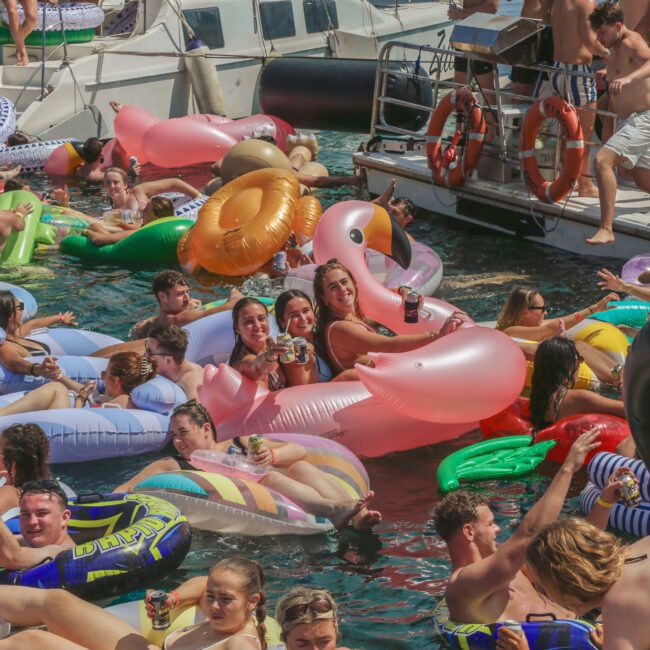 A lively crowd of people relaxes on colorful inflatable pool floats in the water near boats, enjoying a sunny day at a yacht party. Many are smiling, holding drinks, and socializing with friends.