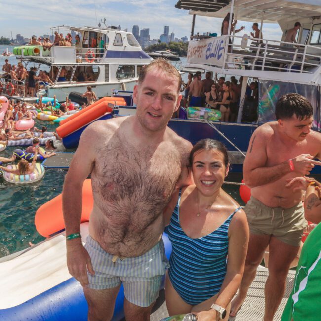 A man and a woman in swimsuits smile at the camera on a boat, surrounded by people, pool floats, and other boats at a lively outdoor party by the water with a city skyline in the background.
