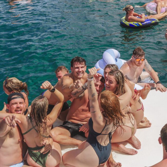 A group of young adults in swimsuits sit closely together on a floating platform, smiling and raising their arms while others relax on inflatable floats in the water around them. The scene is lively and joyful.