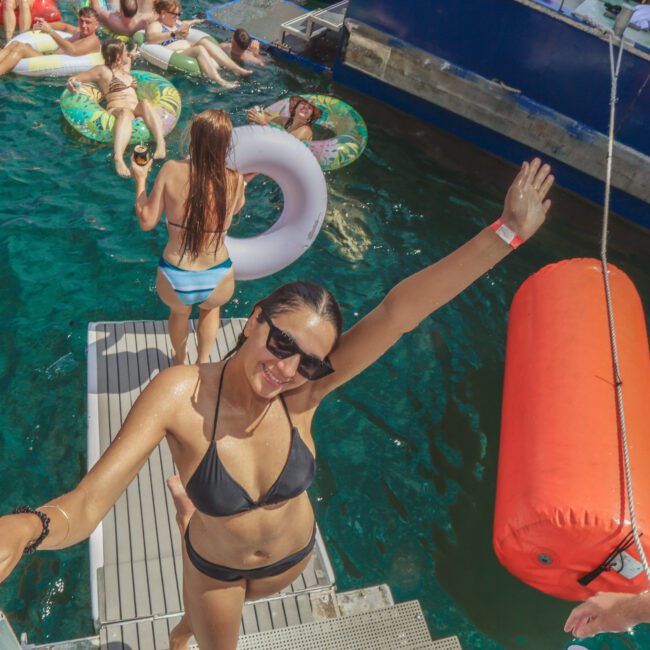 A woman in a black bikini and sunglasses poses smiling on a boat ladder, raising one arm. Behind her, people relax on colorful floaties in clear blue water near a docked boat. The atmosphere is lively and summery.