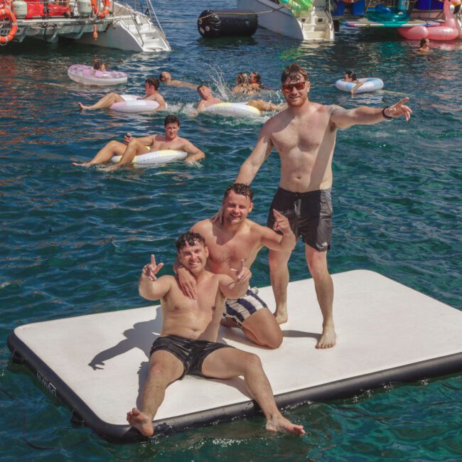 Three men in swim trunks pose and smile on a floating mat in clear blue water, while people relax on floats and yachts are anchored nearby. The scene is sunny and lively, with a party atmosphere.
